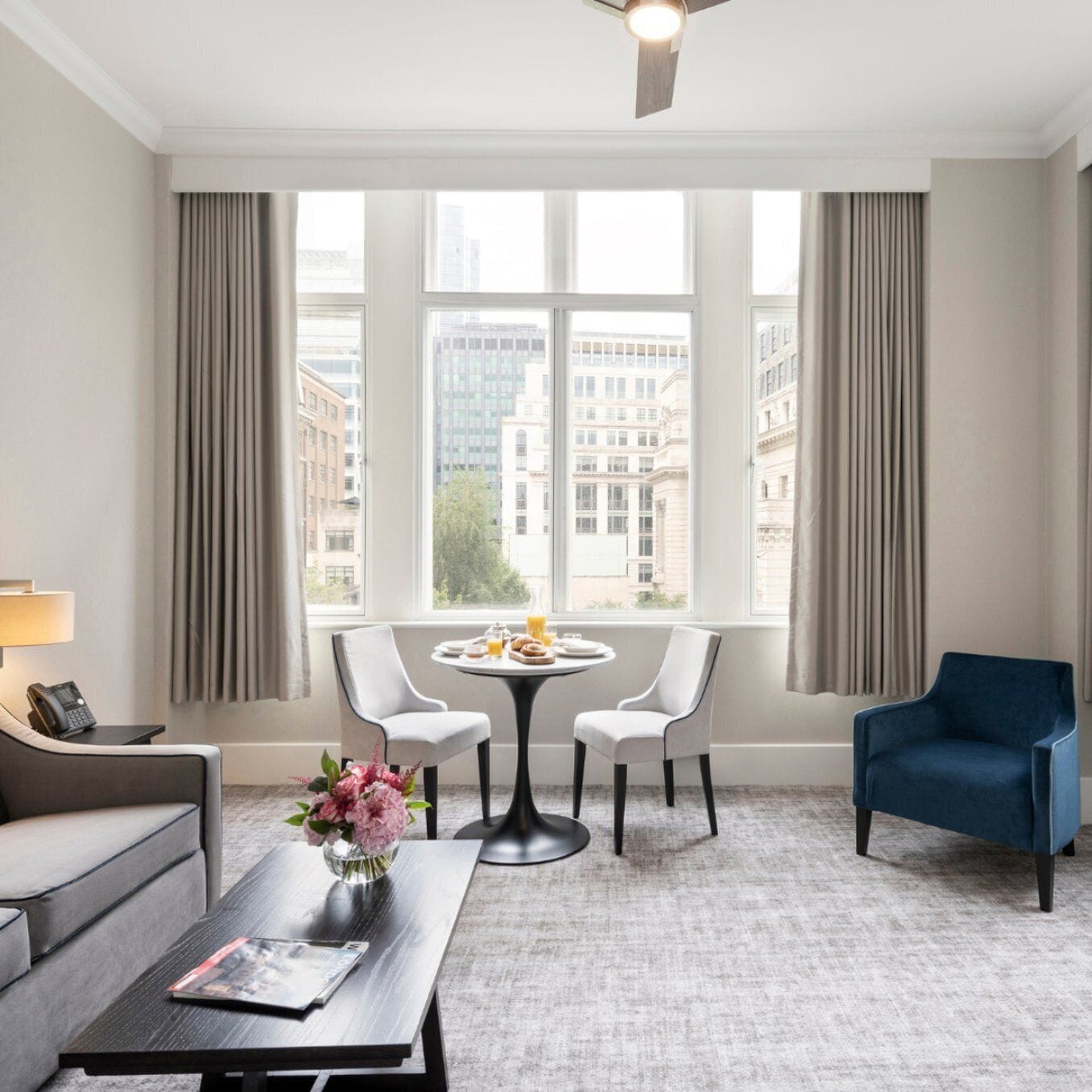 Quarto de hotel com uma grande janela, uma mesa redonda com duas cadeiras, um sofá, uma poltrona azul e uma mesa de centro com um vaso de flores.
