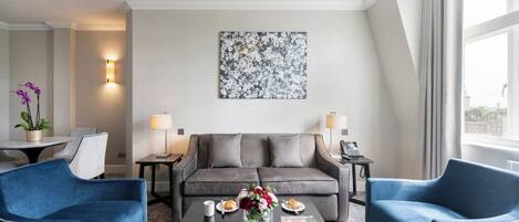 A modern hotel room with a grey sofa, two blue armchairs, a coffee table with flowers, a painting on the wall, and a city view through the window.