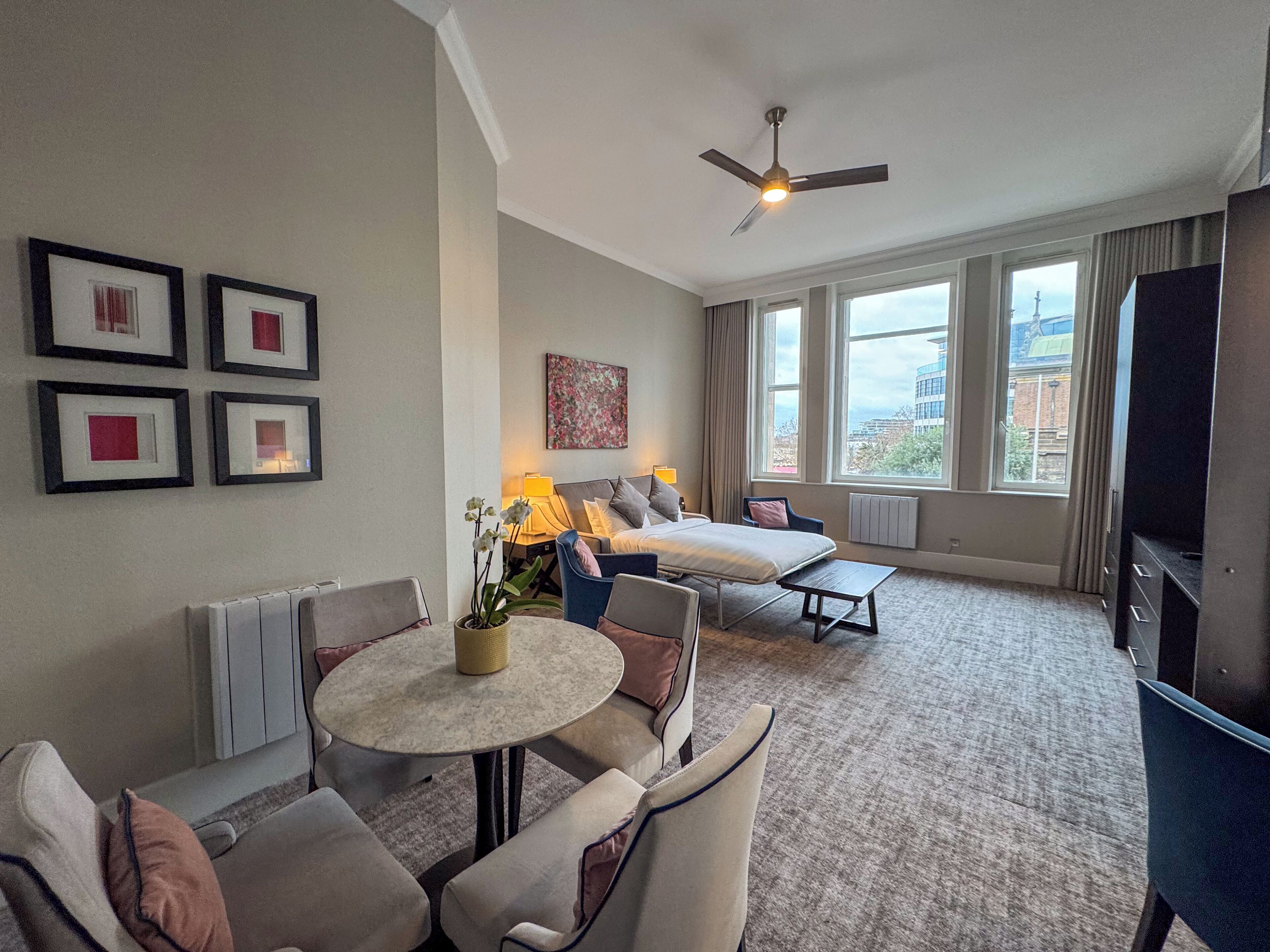 A hotel room with a bed, a sofa, a dining table, and a TV. There are framed pictures on the wall and a window with a view of buildings.