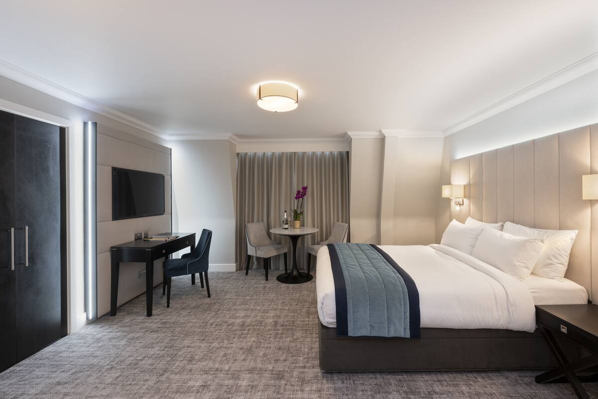 A hotel room with a bed, a desk, a chair, a small table, and a TV.