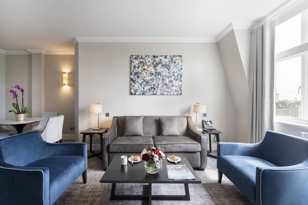 A modern living room with a grey sofa, two blue armchairs, a coffee table with a vase of flowers, and a painting on the wall.
