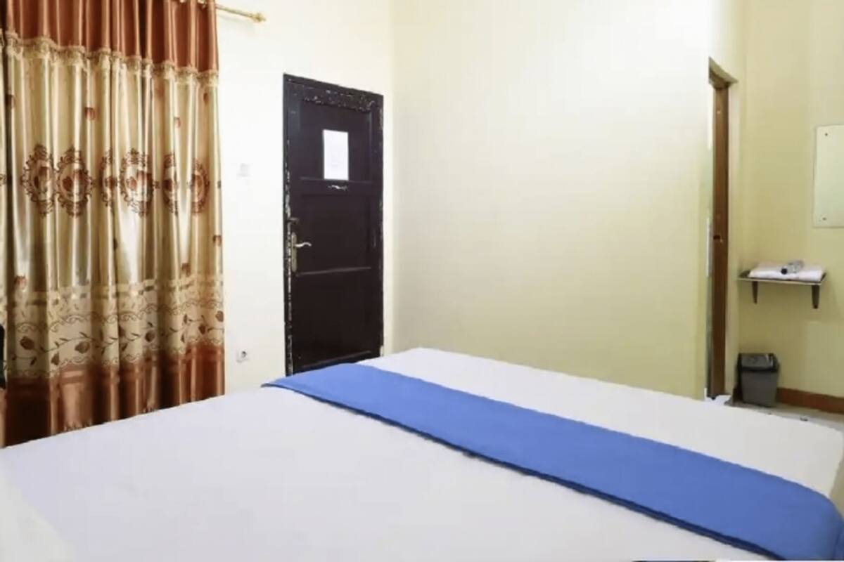A hotel room with a bed, a desk with a computer, a door, and a small shelf with items.