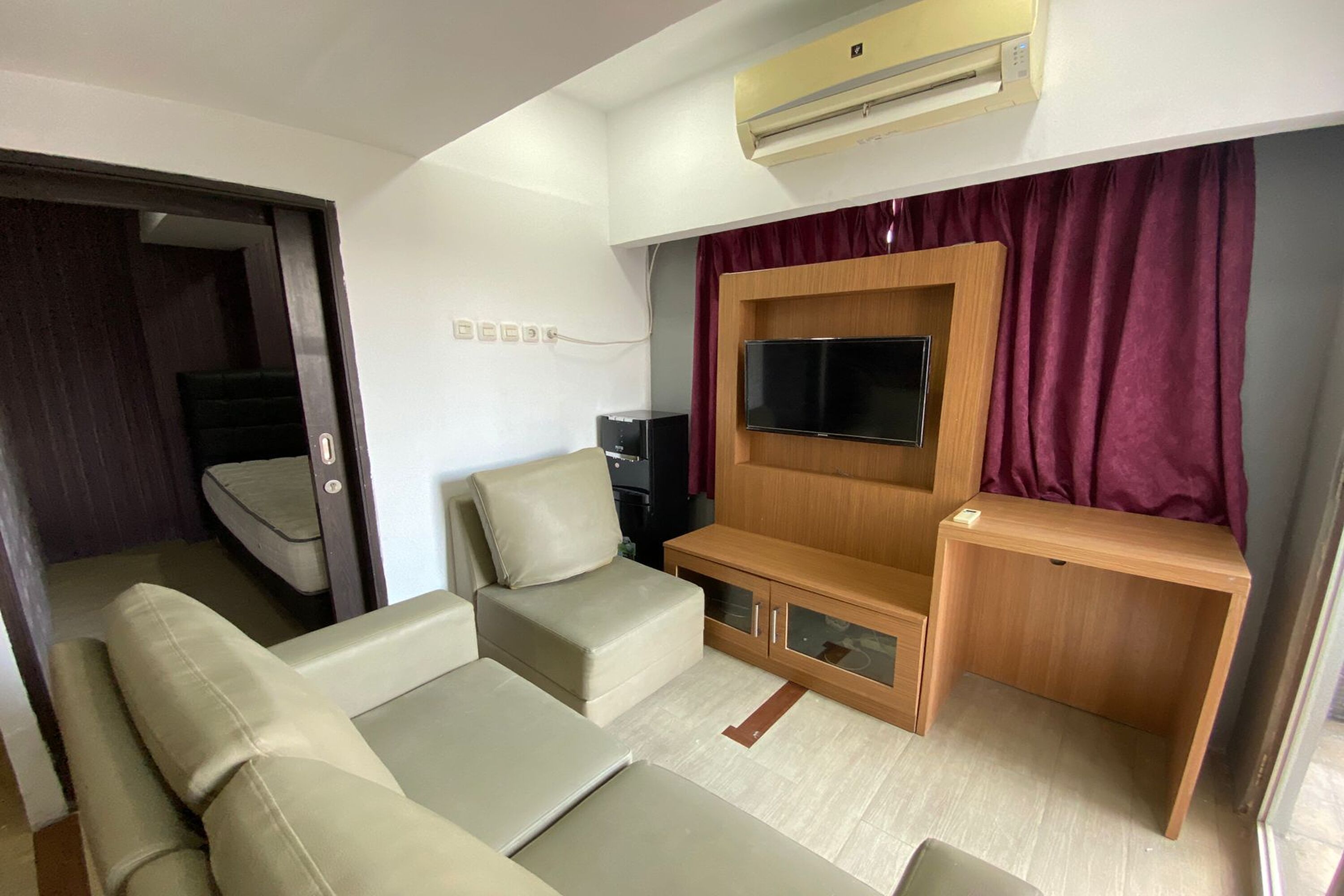 A compact living space with a sofa set, a TV mounted on a wooden cabinet, and a sliding door leading to a dark room.