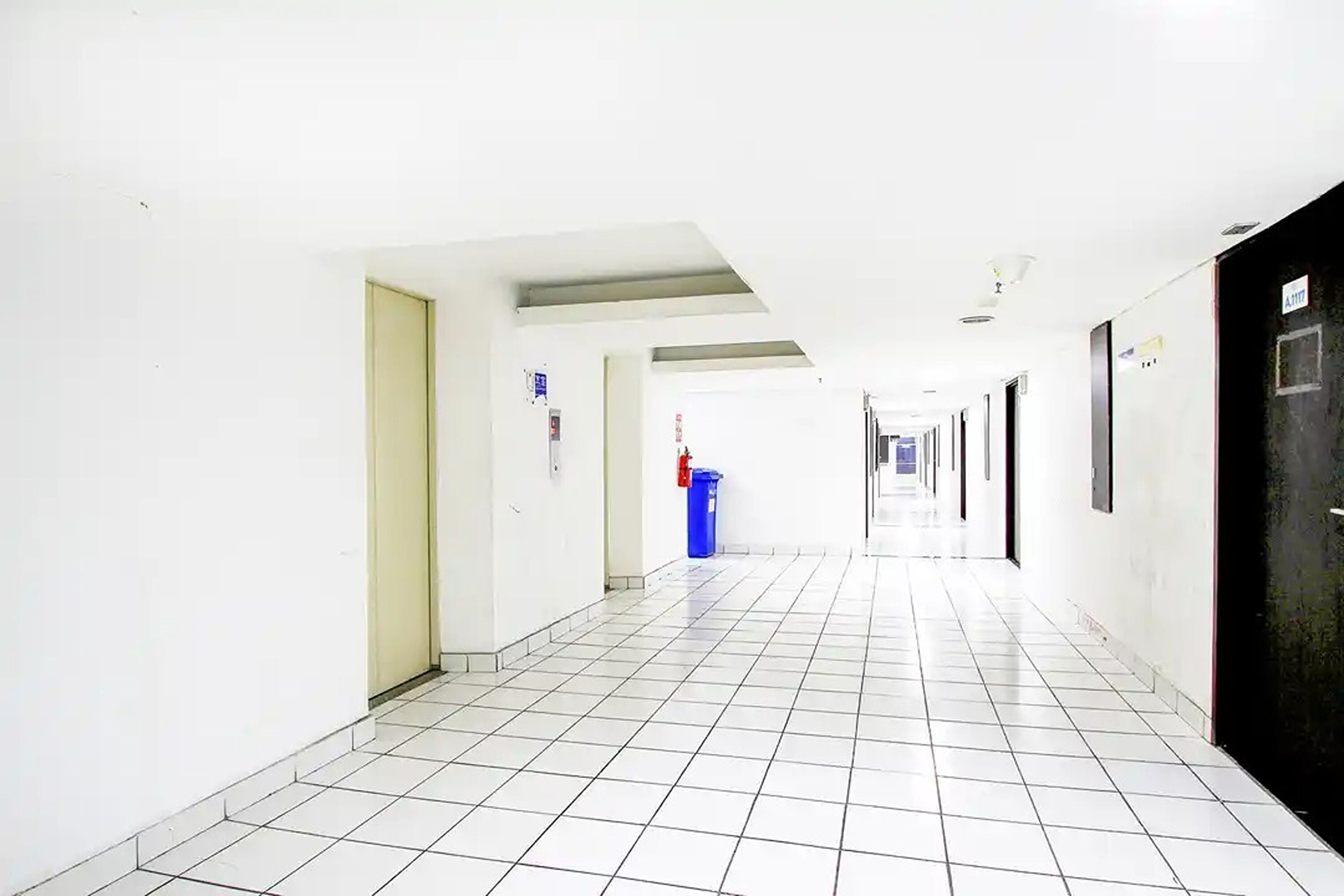 A long, narrow hallway with white tiled floors and walls, doors on both sides, and a blue trash bin in the distance.