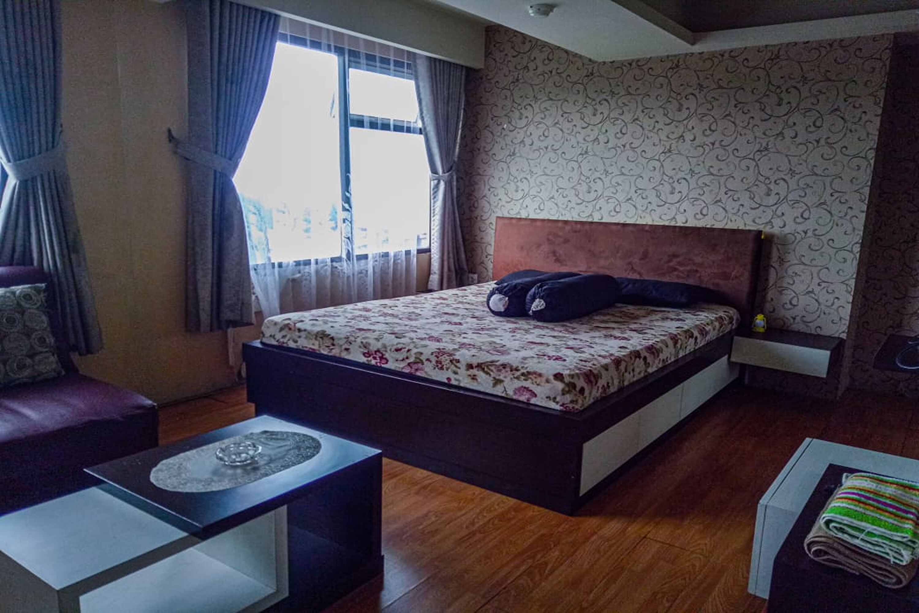 A bedroom with a large bed, a nightstand, a wooden floor, and a window with curtains.