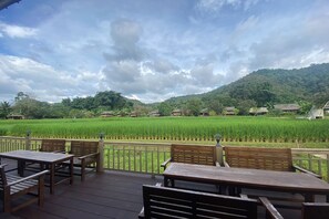 A wooden deck with tables and chairs overlooking a lush green rice field and a mountain in the background. - Lhongkhao Samoeng By Chi Villa (Samoeng)