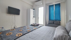 A hotel room with a bed, a TV mounted on the wall, a window with blue curtains, and a built-in wardrobe. - Room Rendi @ Podomoro Golf View (Gunung Putri)