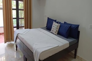 A neatly made bed with a white bedspread and blue pillows, a window with curtains, and a tiled floor.