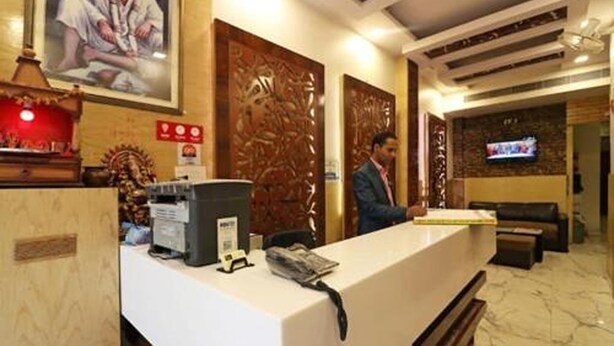 A hotel reception area with a reception desk, a television mounted on the wall, and a seating area with a sofa and armchairs. - Hotel Prem Deluxe Guest House (New Delhi)
