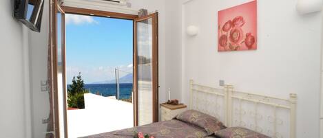A bedroom with a bed, a TV, a painting, and a view of the sea.