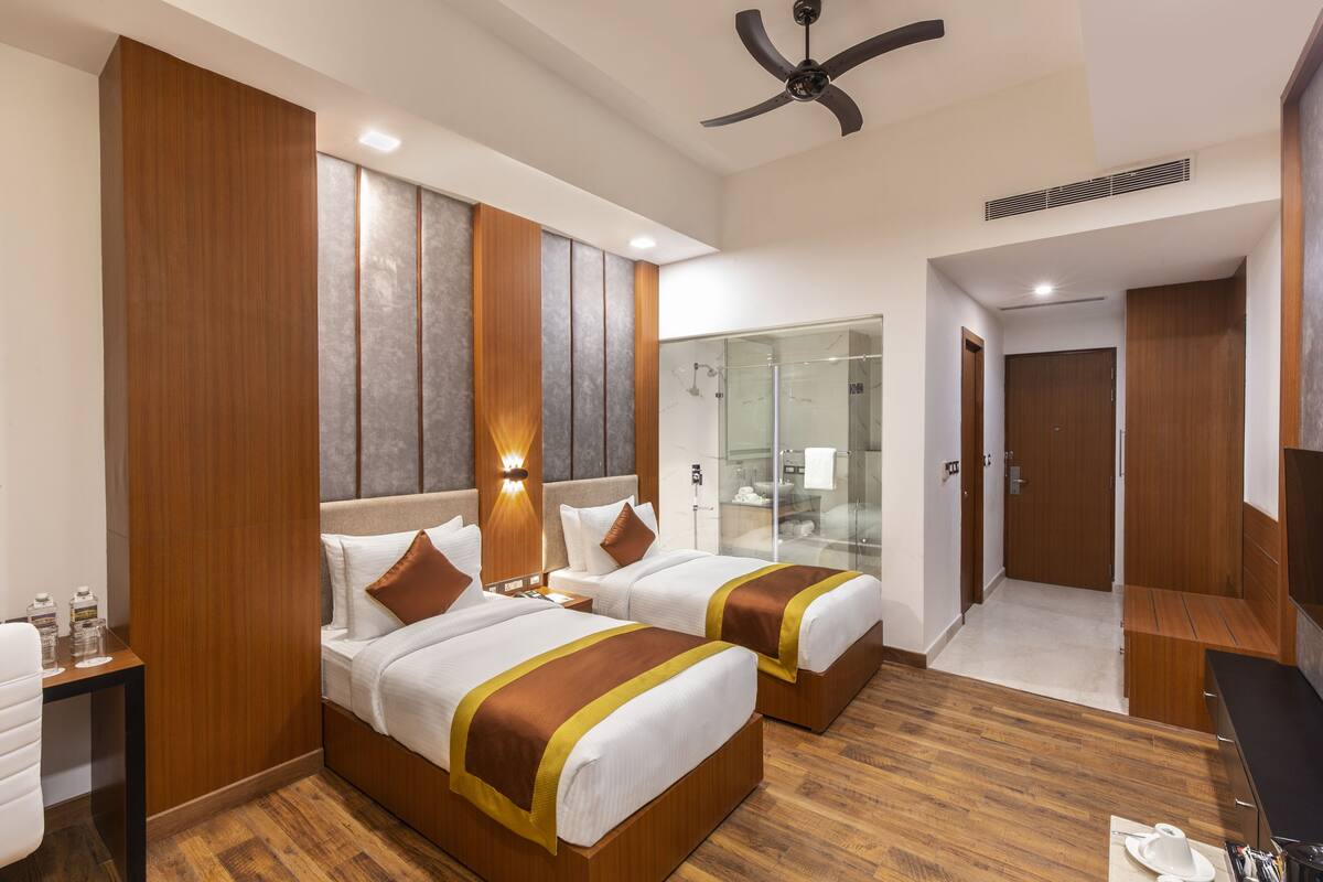 A hotel room with two beds, a desk, a chair, a TV, and a bathroom with a glass shower.