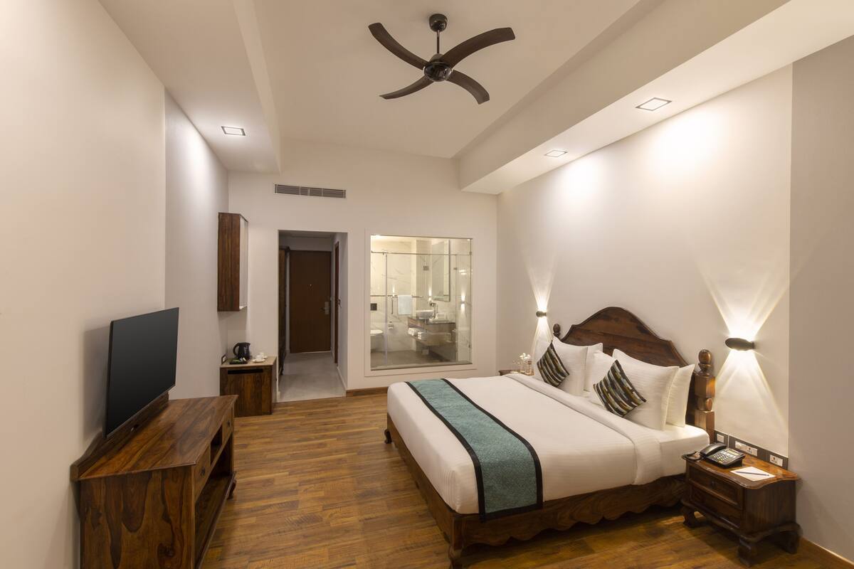 A bedroom with a bed, bedside table, nightstand, TV, ceiling fan, and a bathroom visible through a glass door.