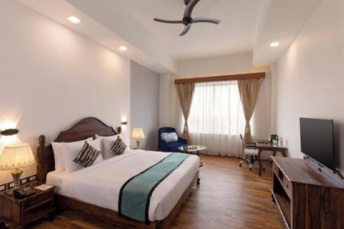 A hotel room with a bed, bedside table, lamp, chair, desk, TV, and curtains.