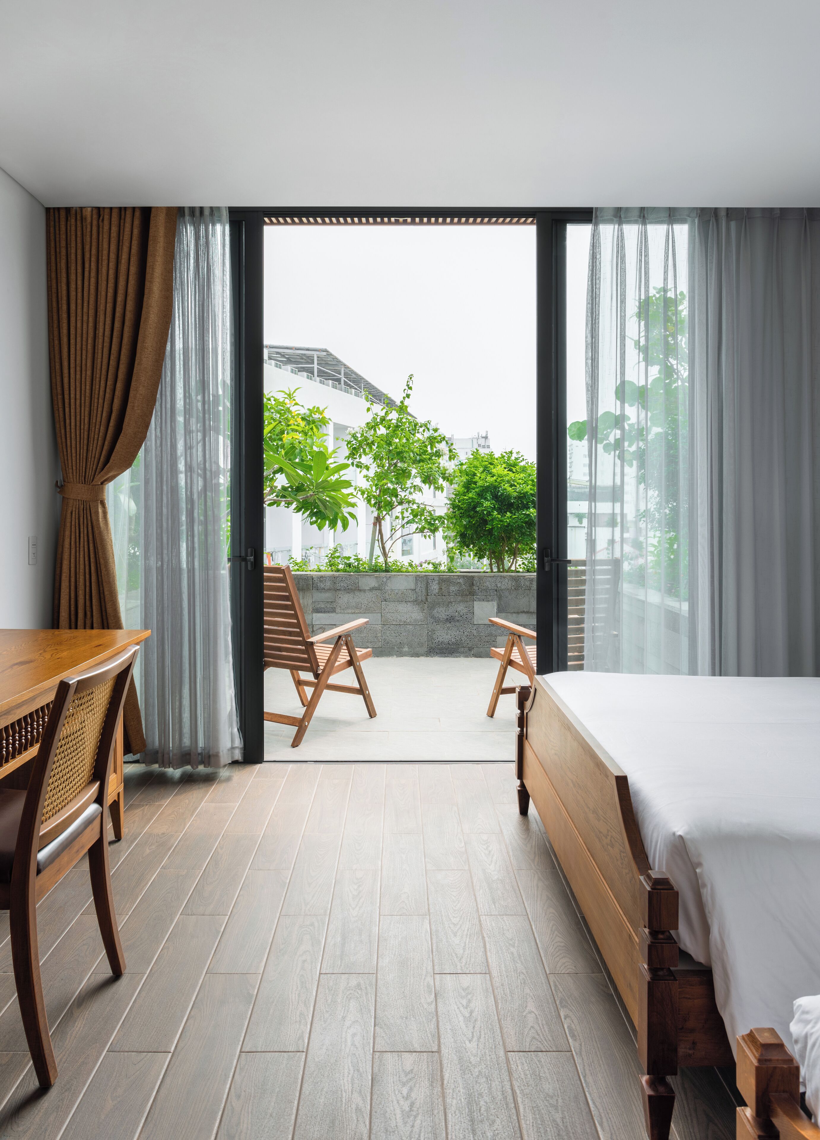 A modern hotel room with a balcony, wooden furniture, and a view of greenery and buildings.