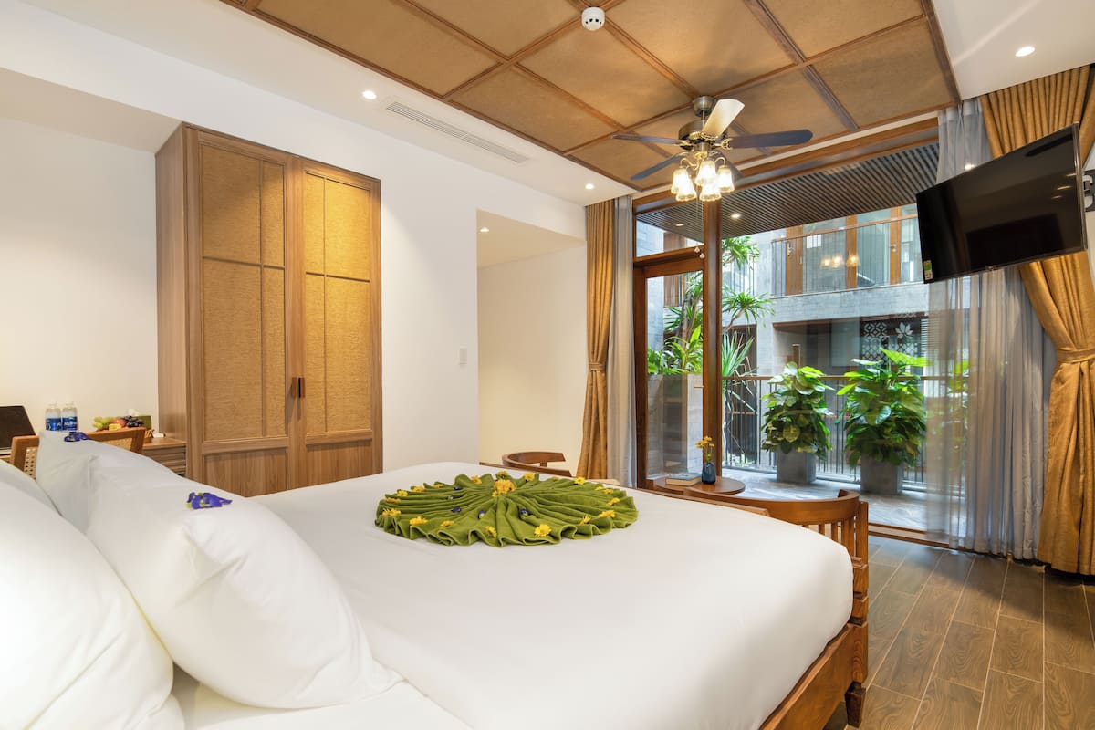 A hotel room with a large bed, a wooden wardrobe, a TV mounted on the wall, and a balcony with potted plants.