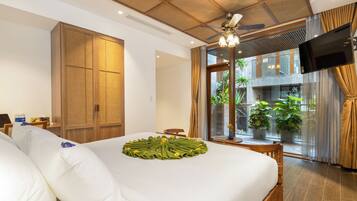 A hotel room with a large bed, a wooden wardrobe, a TV mounted on the wall, and a balcony with potted plants.