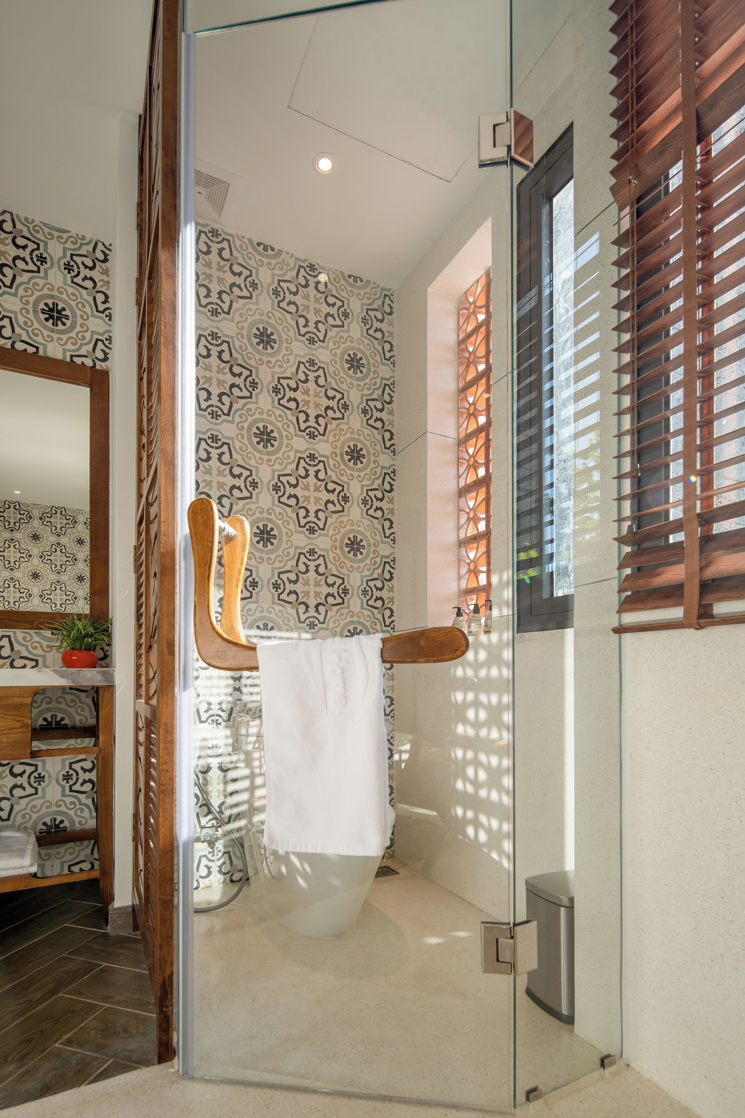 A bathroom with patterned tiles, a glass shower enclosure, wooden blinds, and a white towel hanging on a rack.