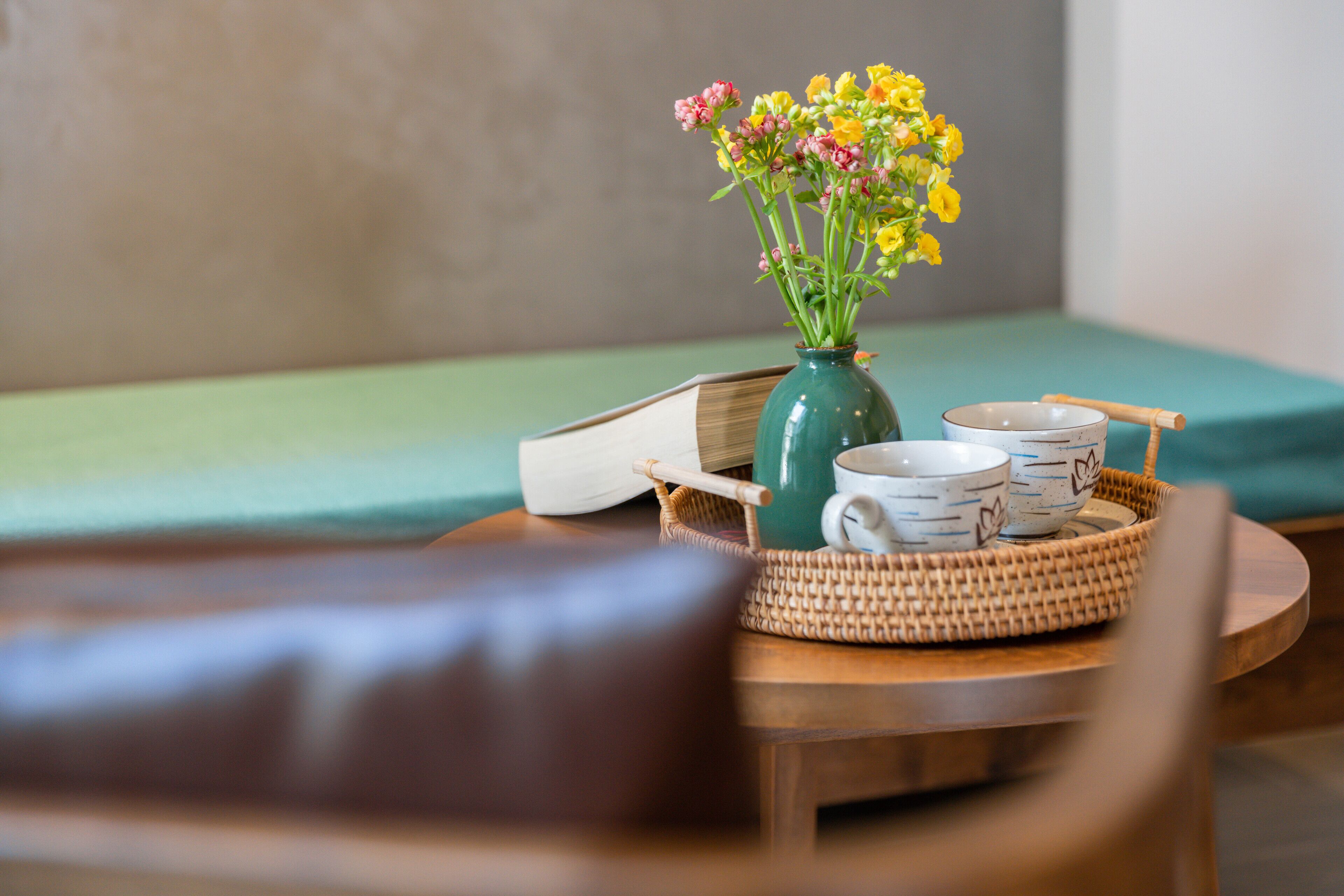 A wooden table with a woven tray holding a green vase with yellow flowers, two white cups with blue patterns, and a book.