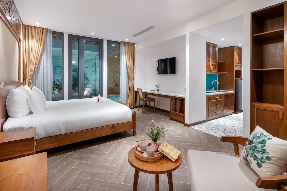 A modern hotel room with a large bed, a desk, a TV, and a kitchen area.