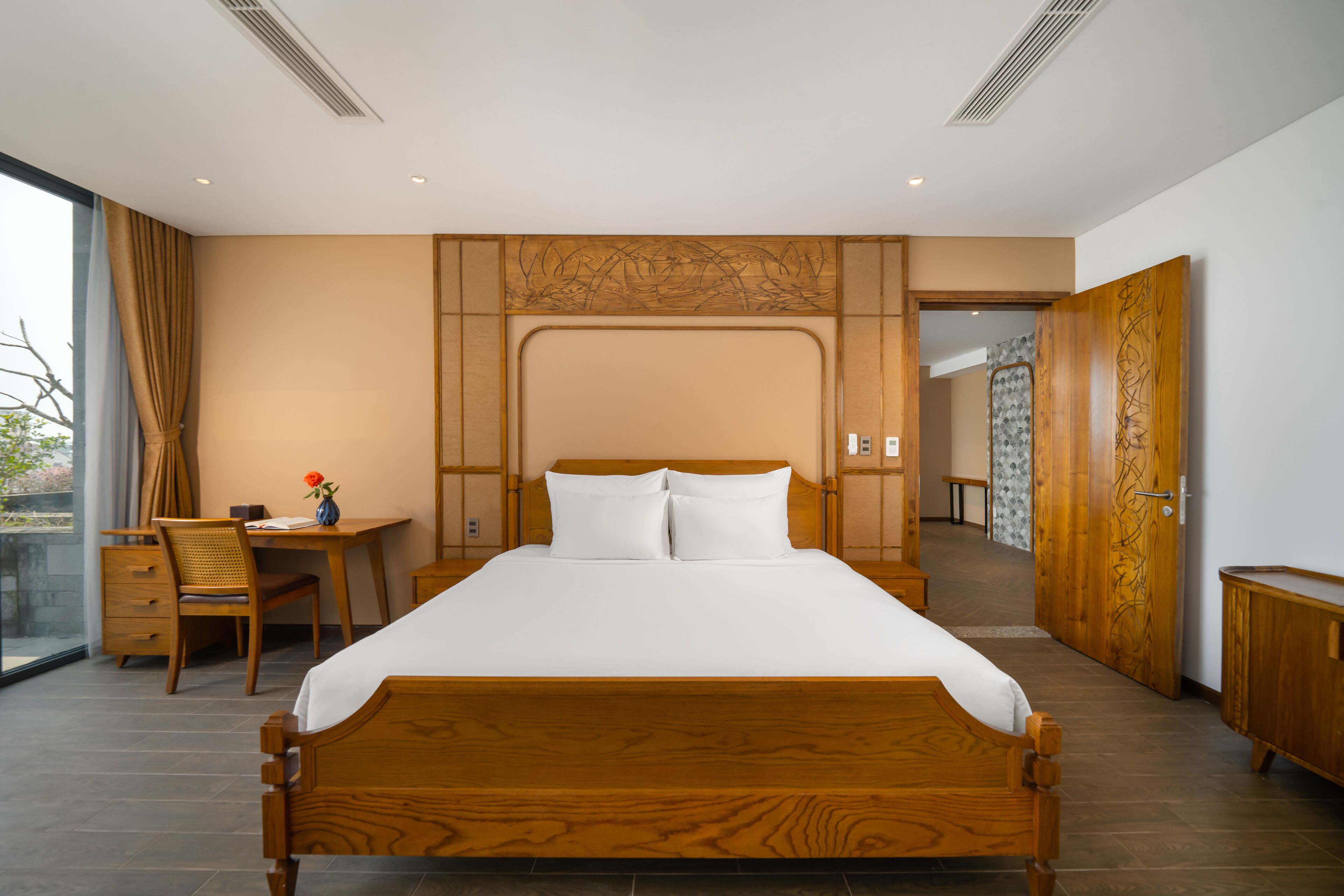 A modern hotel room with a large bed, wooden headboard, a desk with a chair, and a view through a large window.