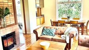 A living room with a fireplace, wooden flooring, a sofa, a coffee table, and a dining area with a window view. - Prime Spot! 5mins to “Grand Cliff Top Walk” & “3 Sisters” (Katoomba)