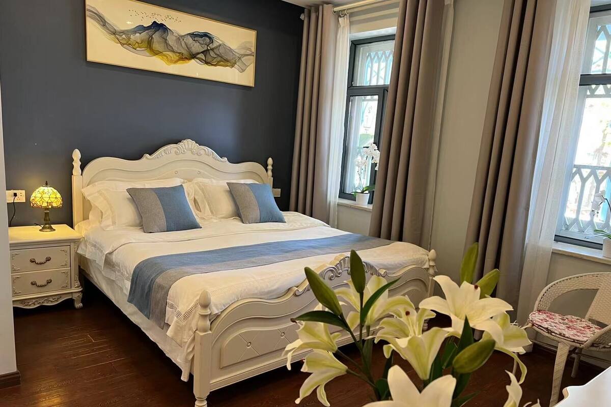 A bedroom with a white bed, a nightstand with a lamp, a window with curtains, and a painting on the wall.