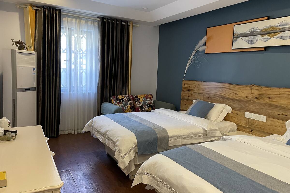 A hotel room with two beds, a desk, a window with curtains, a wall-mounted painting, and a wooden headboard.