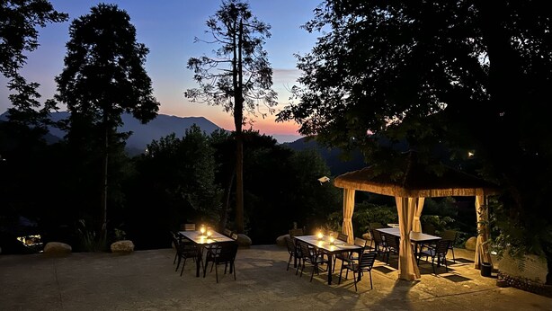 Un área de comedor al aire libre con mesas y sillas, un pabellón con techo de paja y montañas al fondo al anochecer.