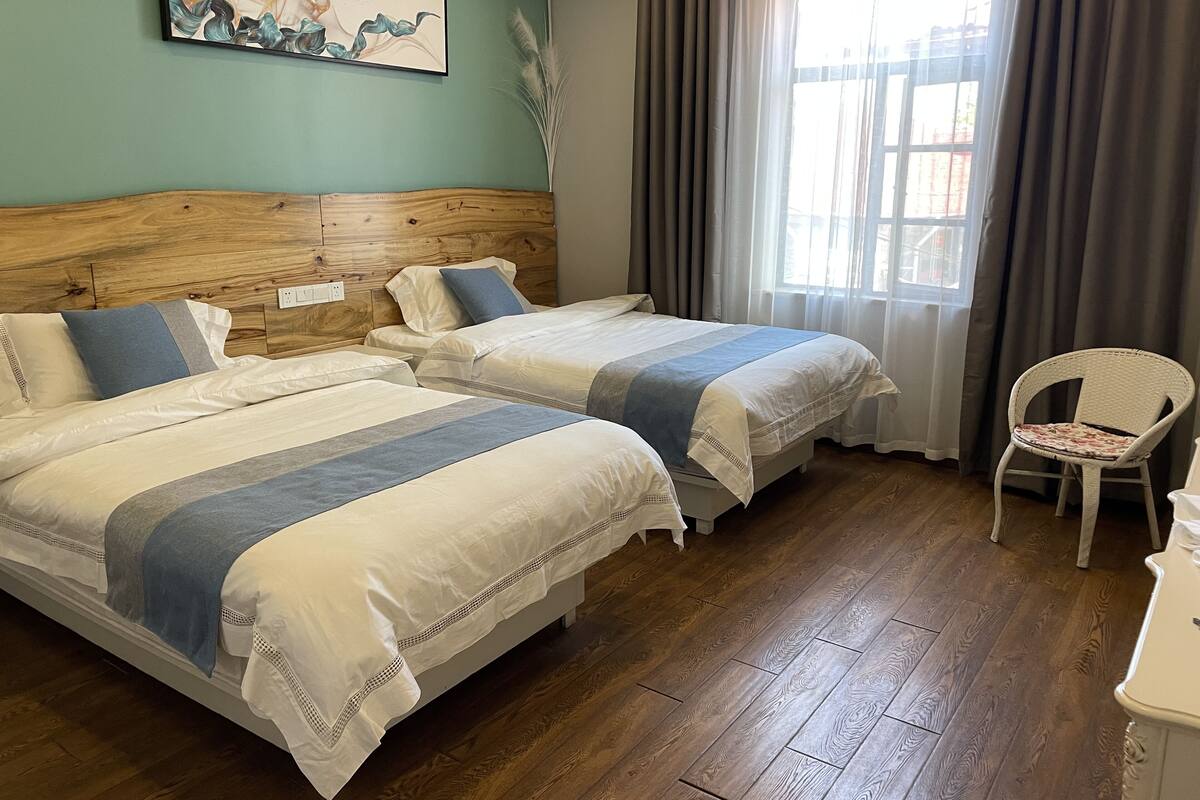 A hotel room with two beds, a wooden headboard, a window with curtains, a chair, and a painting on the wall.