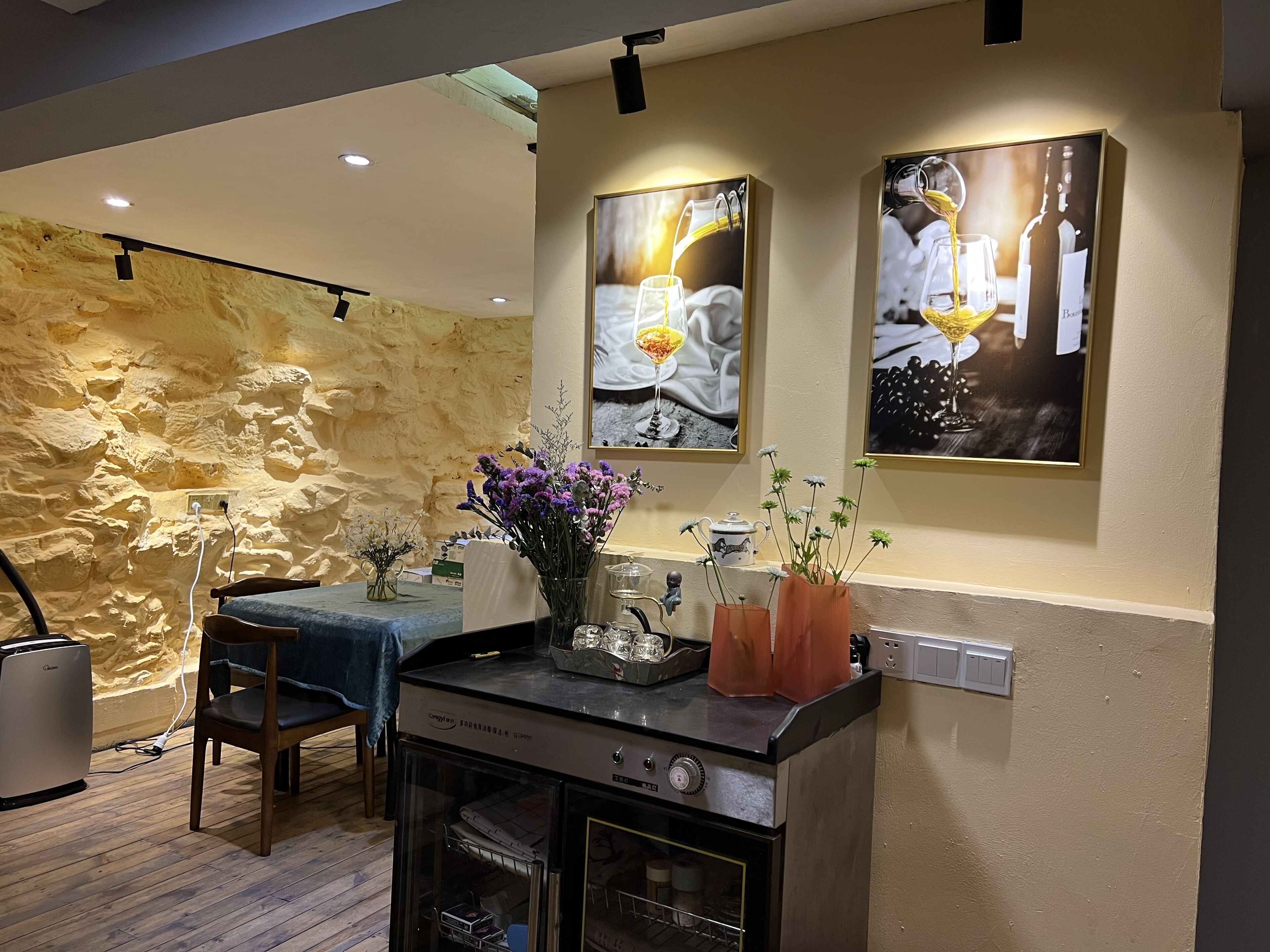 A kitchen with a stone wall, a dining table with chairs, a coffee machine, and two framed pictures on the wall.