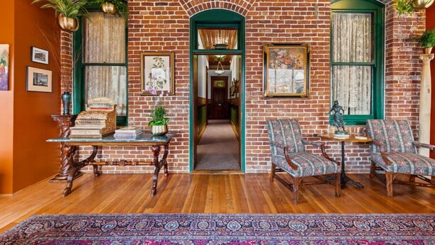A room with brick walls, wooden flooring, and a green archway leading to another room. There are two chairs, a table with books and a plant, and framed pictures on the walls.