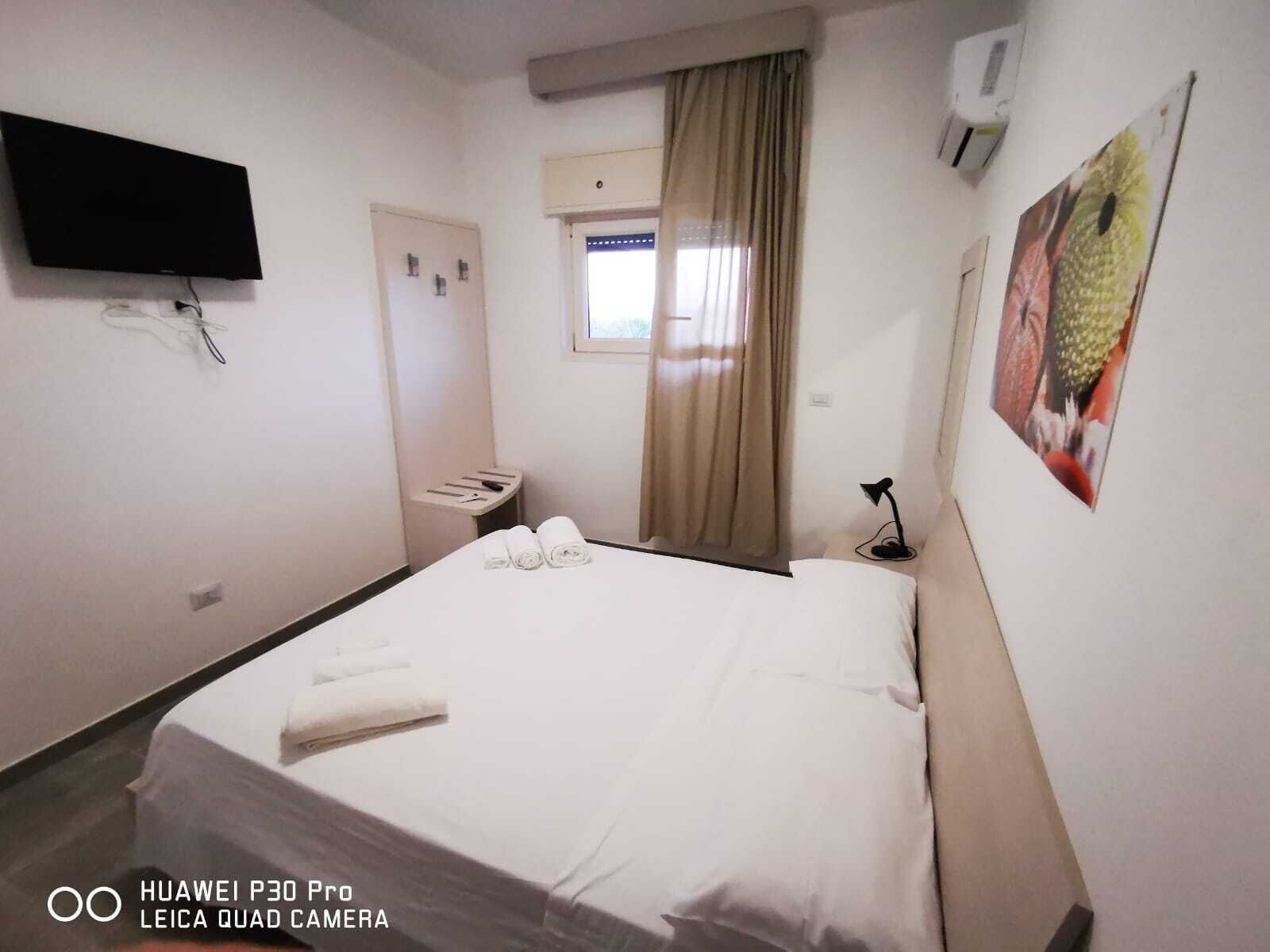 A hotel room with a bed, a flat-screen TV, a window with curtains, a wall-mounted air conditioner, and a small table with a lamp.