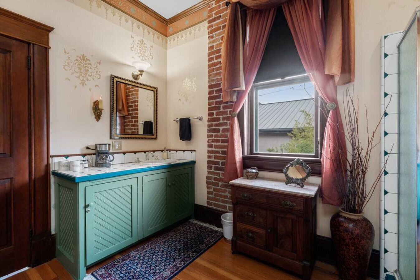 A bathroom with a green vanity, a mirror, a window with curtains, and a vase with branches.