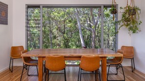 A dining area with a large wooden table and four chairs, a view of a forest through a large window, and hanging plants.