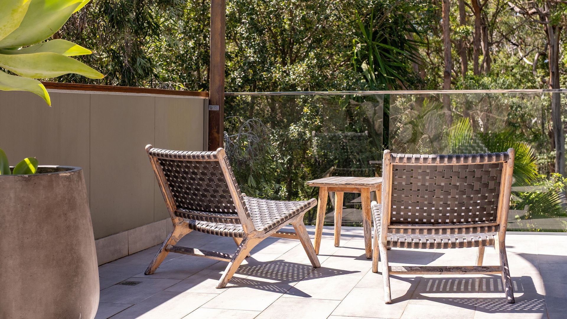 Two wicker chairs with a small wooden table, set on a tiled patio with a view of greenery.
