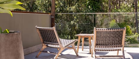 Dos sillas de mimbre con una pequeña mesa de madera, ubicadas en un patio con baldosas y vistas a zonas verdes.