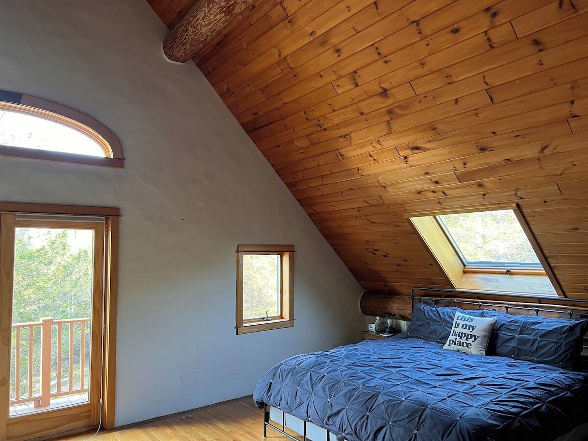 A bedroom with a sloped ceiling, a bed with blue bedding, two windows, and a balcony door.