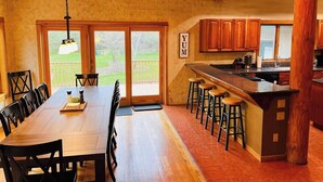 Une cuisine spacieuse avec un îlot central en bois, de grandes fenêtres et un coin repas comprenant une longue table et des chaises.