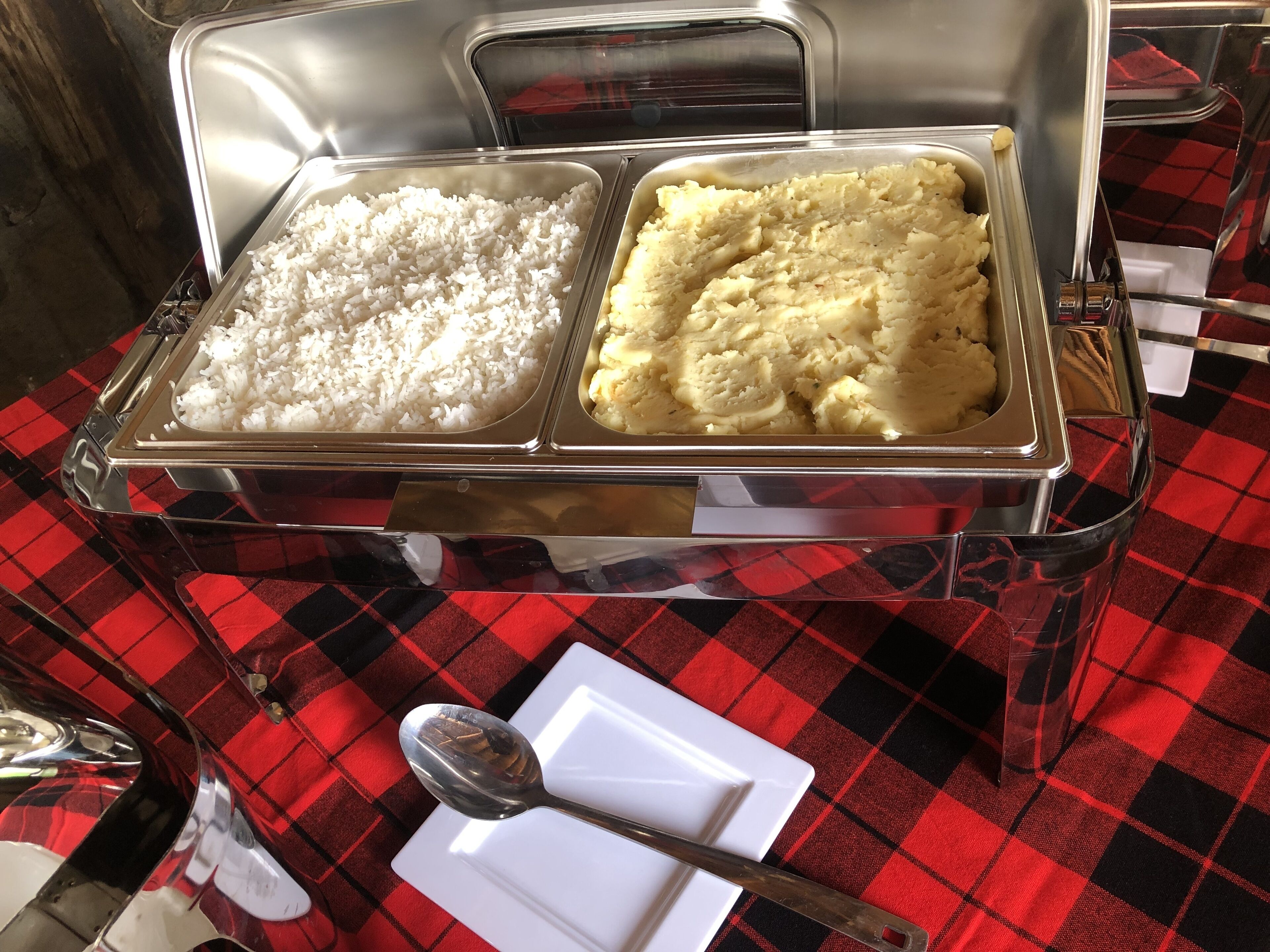 Un bufé con dos fuentes de comida, una de arroz y otra de puré de papas, sobre un mantel a cuadros rojo y negro.
