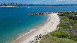 A coastal beach with clear waters, a rocky breakwater, and a forested area.