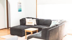 Ein modernes Wohnzimmer mit einem grauen Ecksofa, einem Holztisch und einem kleinen Hocker. An der Wand hängt ein Schild mit Strandmotiven.