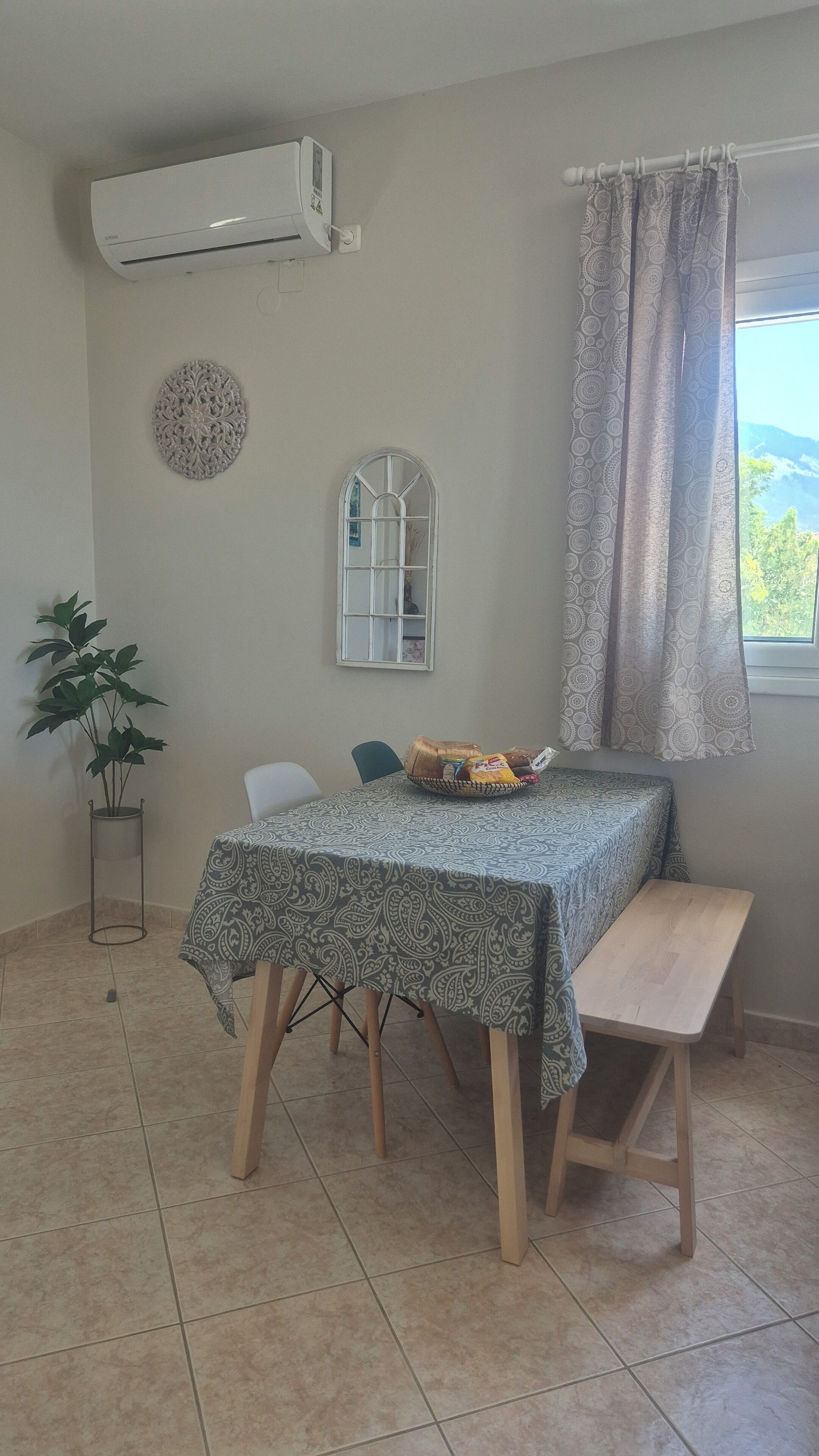 Uma área de refeições com uma mesa coberta por uma toalha estampada, um banco, uma planta em vaso e uma janela com cortinas padronadas.