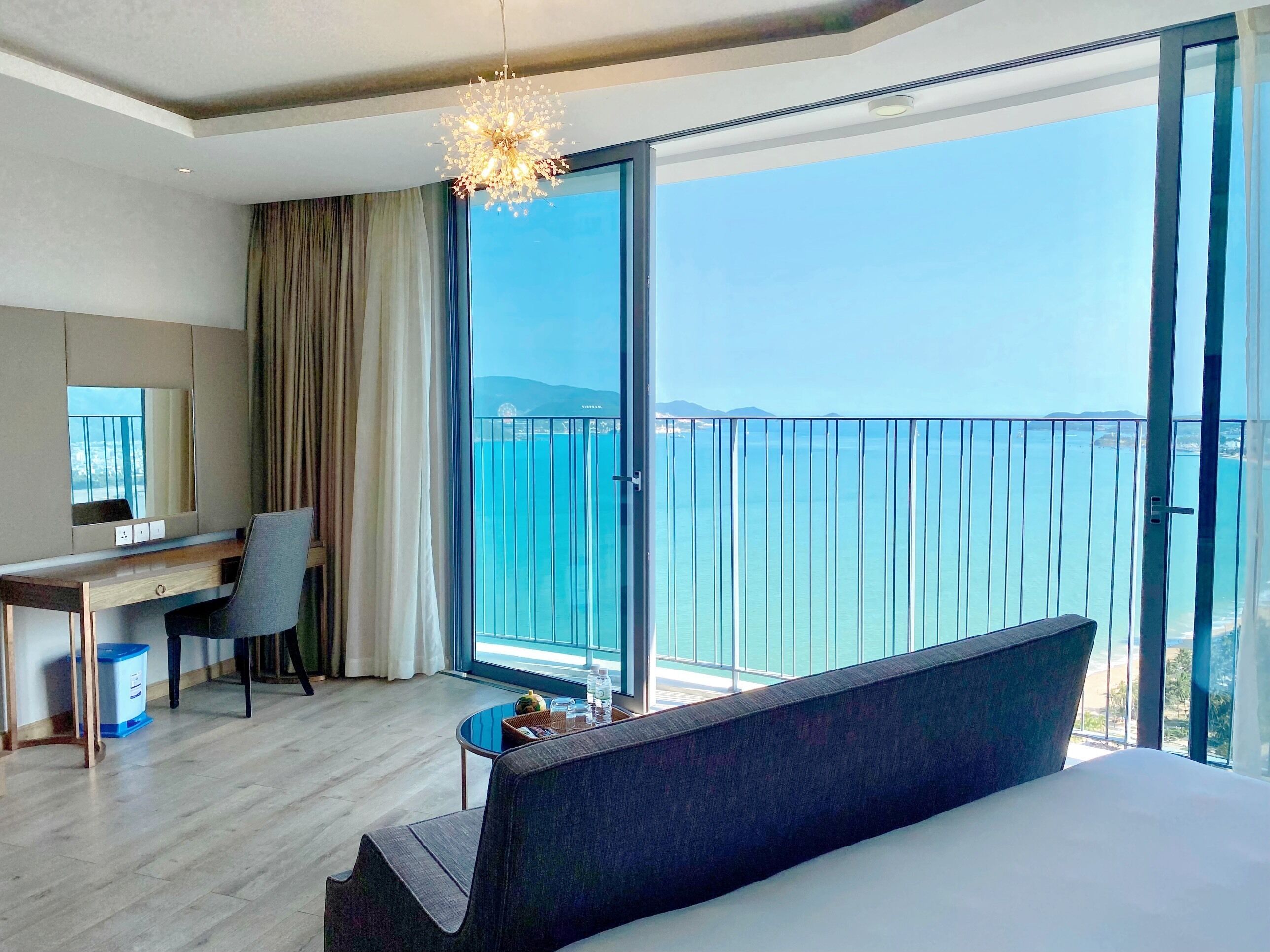A modern hotel room with a large bed, a desk, and a chair. There is a balcony with a view of the sea.