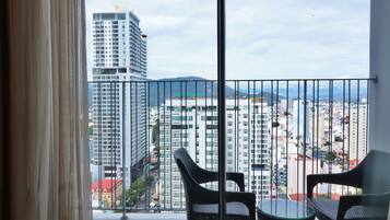 A balcony with two wicker chairs and a glass table, overlooking a cityscape with high-rise buildings and mountains in the distance.