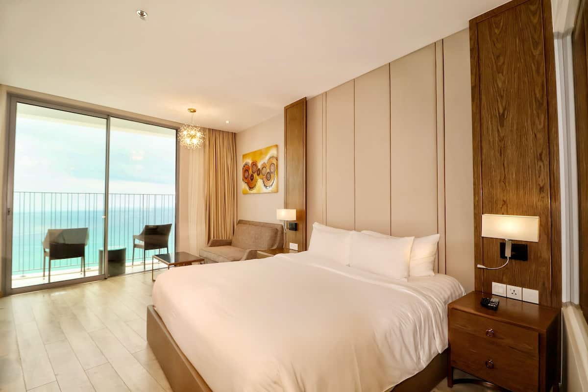 A hotel room with a large bed, a desk, a chair, a nightstand, and a view of the ocean through a sliding door.
