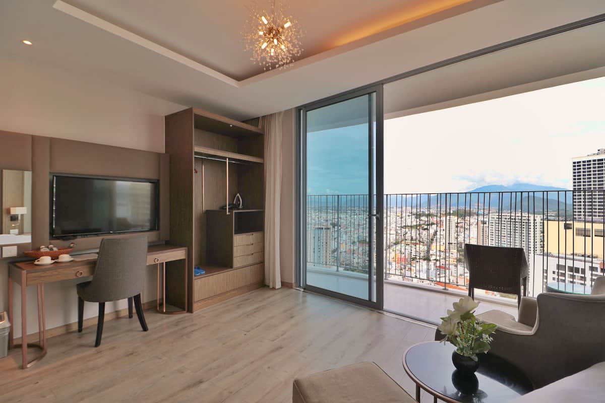 A modern hotel room with a large balcony, a desk with a TV, and a comfortable seating area.