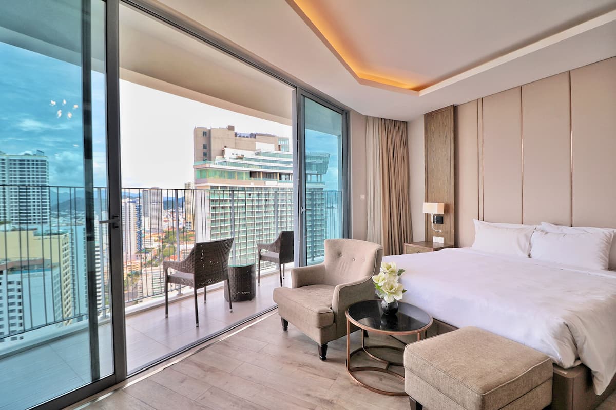 A modern hotel room with a large bed, a sitting area with a chair and a small table, and a balcony with a view of the cityscape.