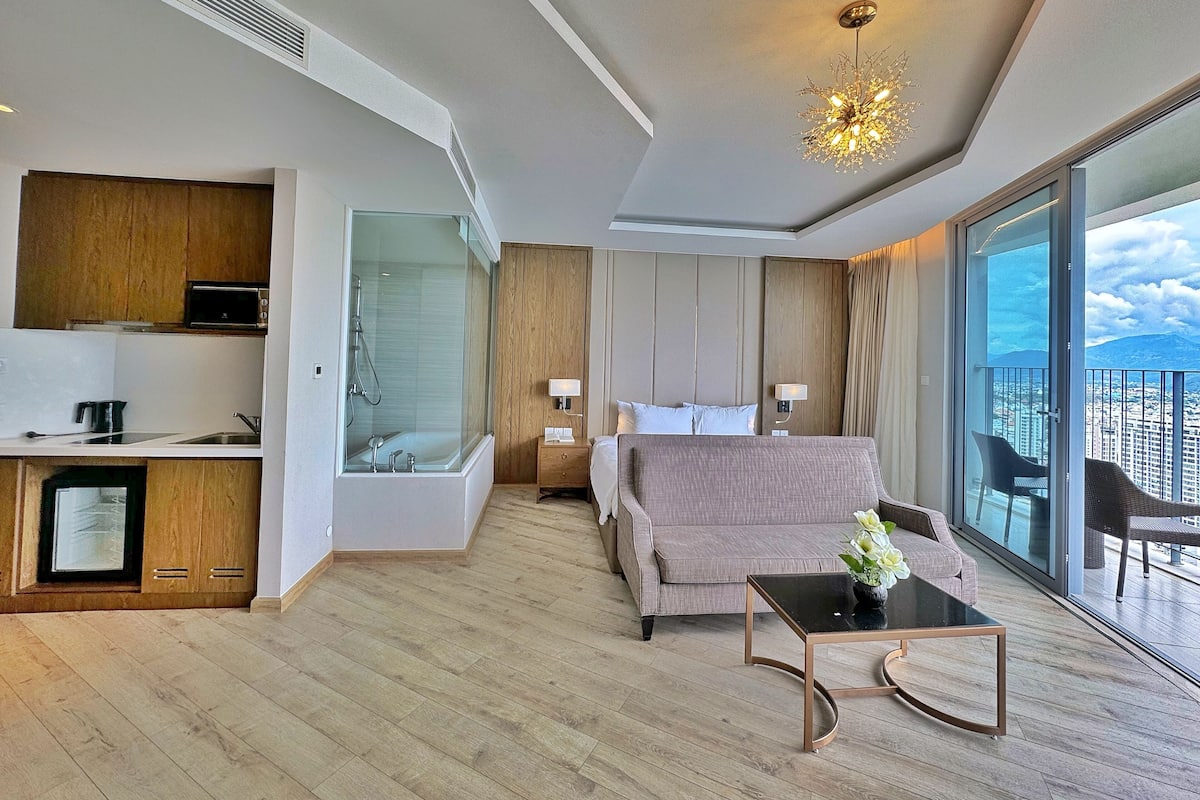 A modern hotel room with a kitchenette, a shower, a sofa, a small table, and a balcony with a city view.
