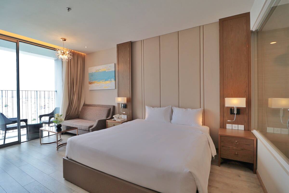 A modern hotel room with a large bed, a sitting area with a chair and table, a balcony with a view, and a painting on the wall.