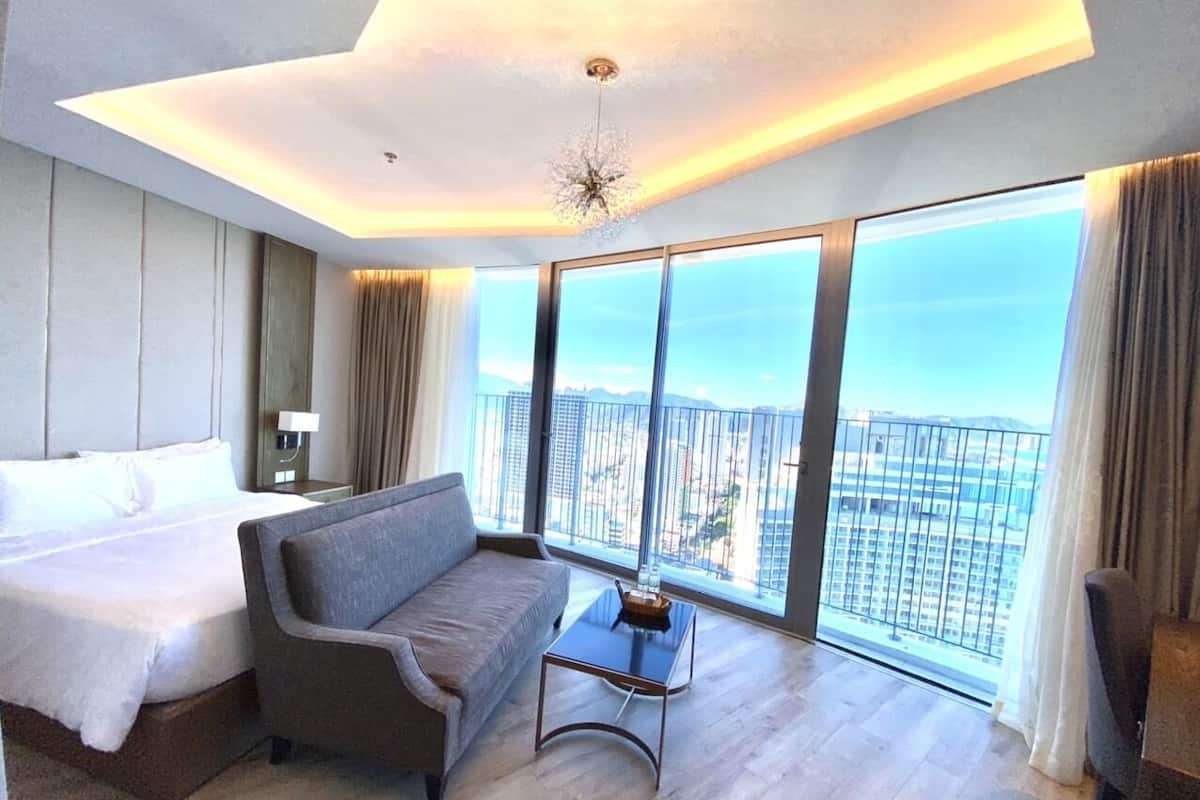 A modern hotel room with a large bed, a sofa, a small table, and a view of the city through large windows.